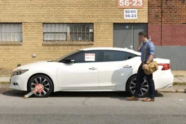 A man with a brown satchel walks past a white sedan parked on the street, which has a yellow and red Risetek Self Releasing Boot locked onto its front left wheel. The car is in front of a brick building with a window and a sign reading 