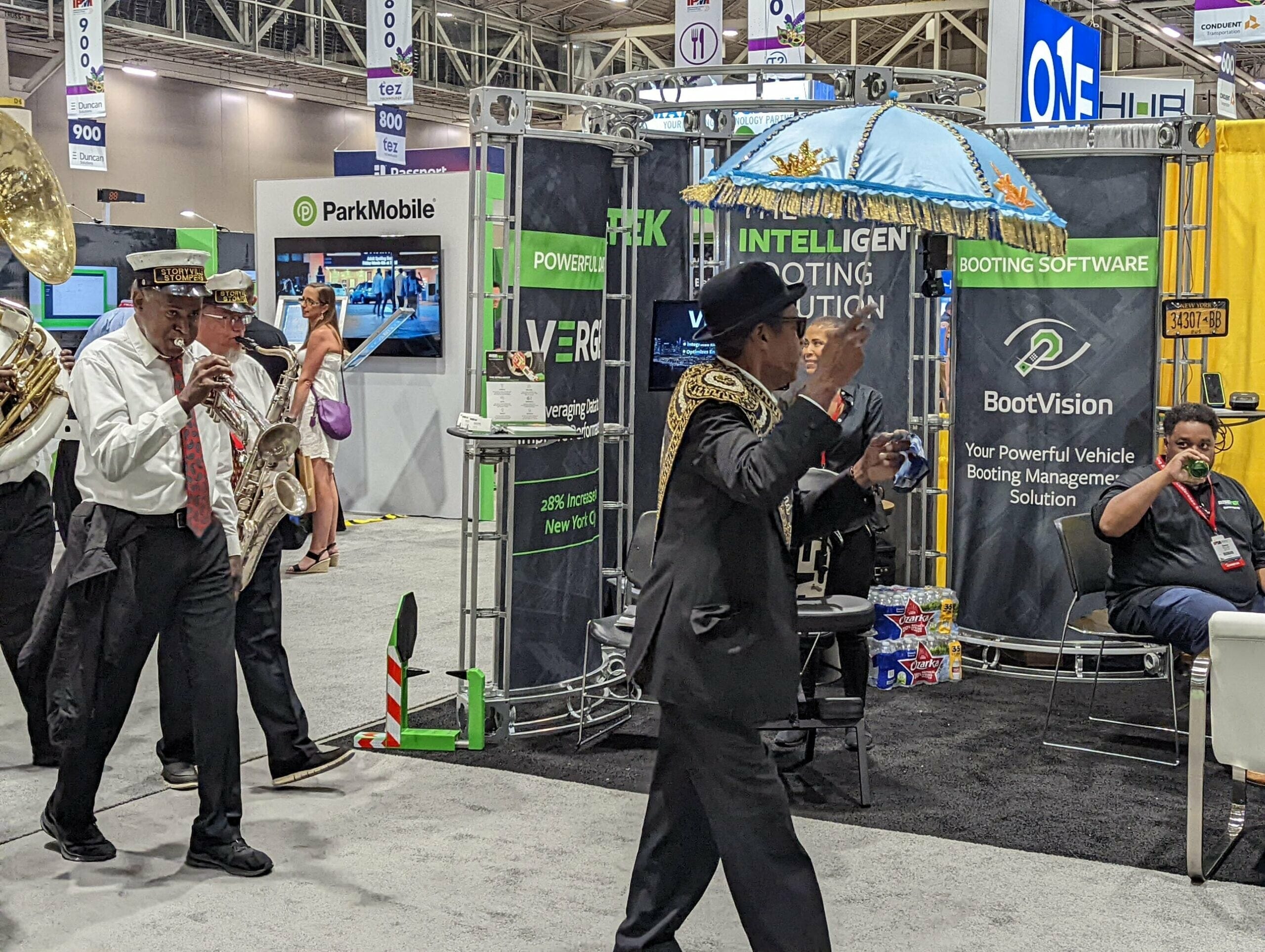 A jazz band in black and white attire, one holding a decorated umbrella, plays instruments while walking past a BoothVision vehicle software display at the Apartmentalize Expo. A seated man watches as they perform inside the busy NAA 2022 convention hall.