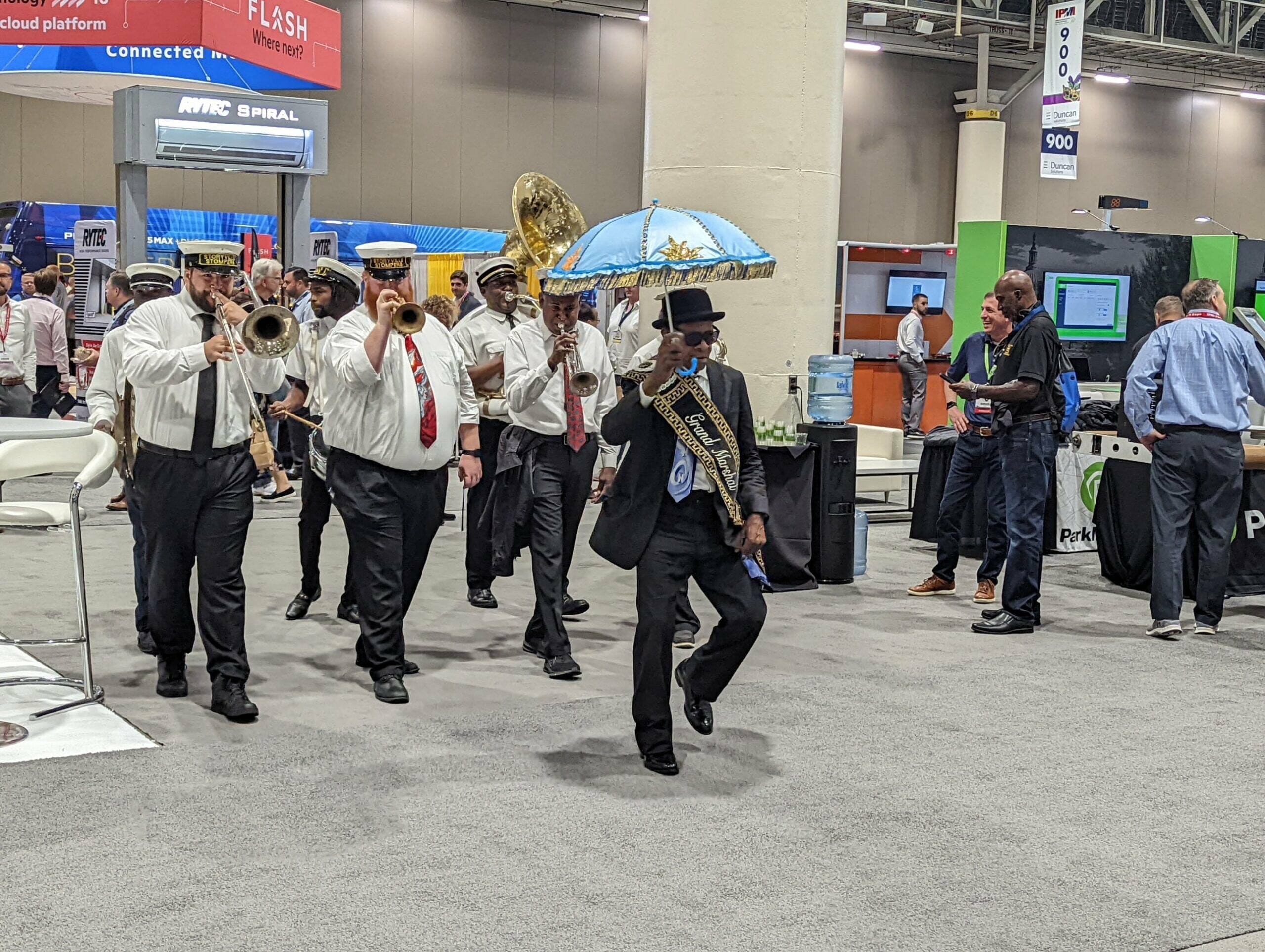 A small brass band in white shirts and ties marches indoors behind a man in a sash and top hat holding a blue parasol at the 2022 NAA Apartmentalize Expo. Attendees, including RISETEK Global staff, watch from exhibit booths in this lively, celebratory scene.