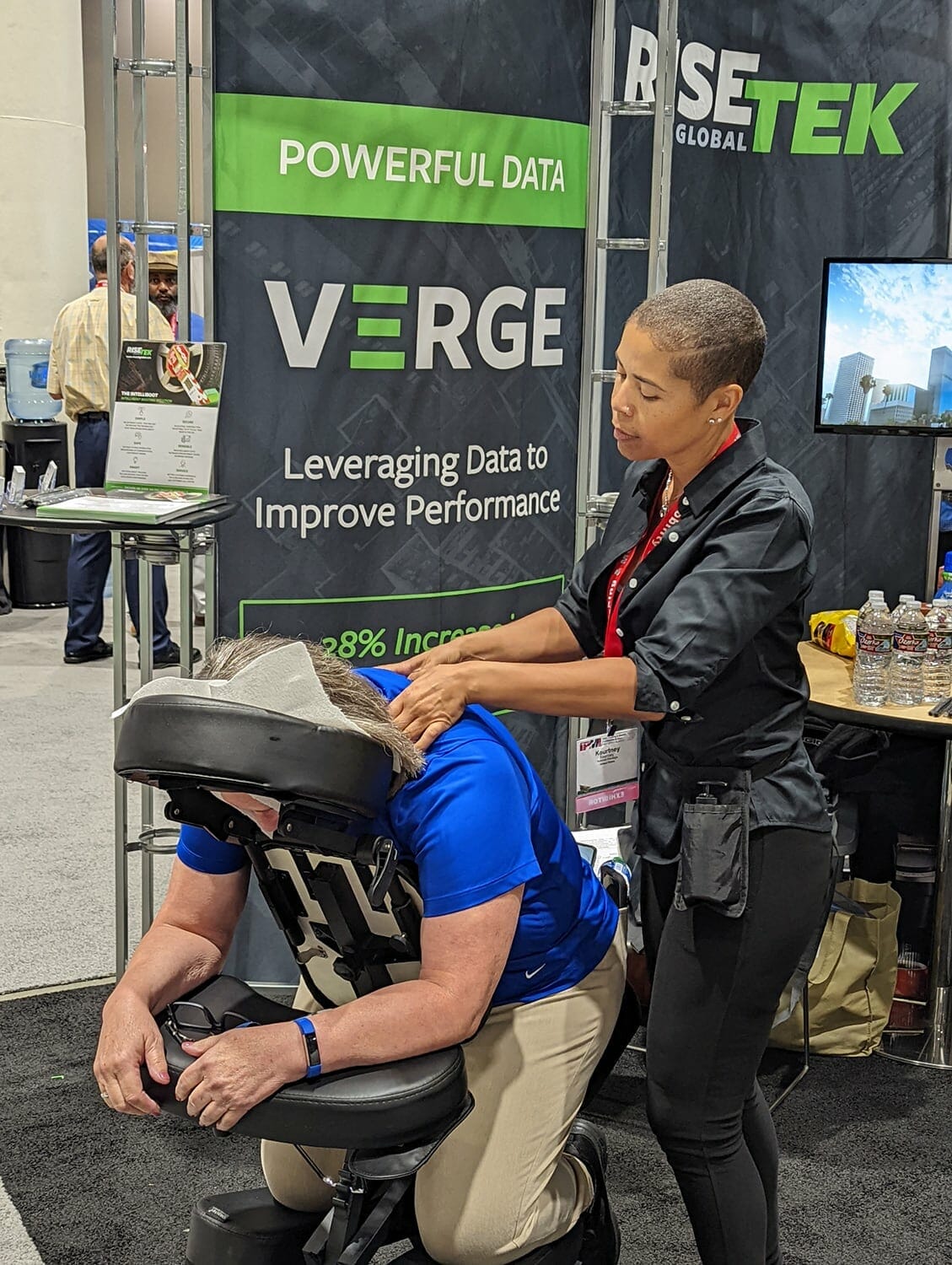 At the NAA Apartmentalize Expo 2022, a woman gives a seated shoulder and neck massage to another woman at the RISETEK Global Exhibits booth. Banners displaying “POWERFUL DATA” and event giveaways are visible in the background.