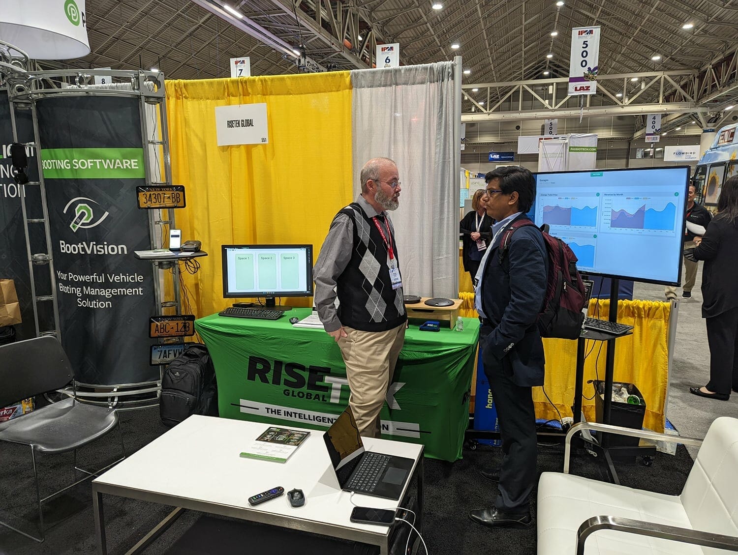 Two men in business attire talk at a trade show booth at IPMI 2022, featuring yellow and white drapes, green 